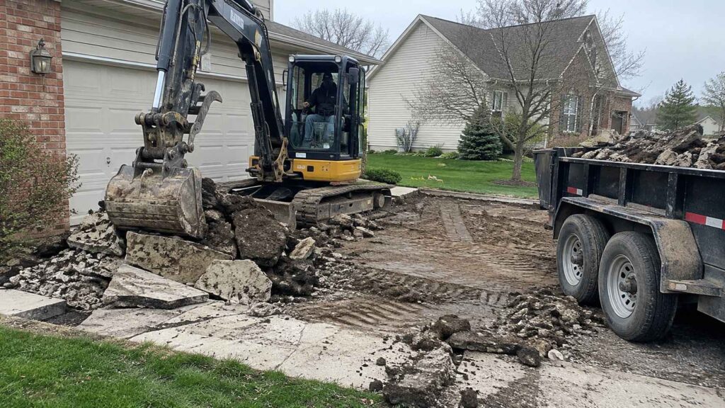 Mini excavator breaking up an old concrete driveway beside a residential garage, with debris loaded into a trailer.